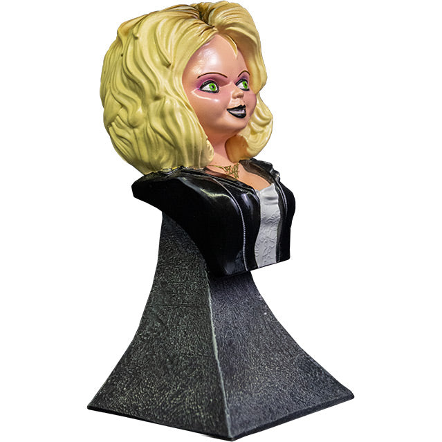 Right side view. Tiffany mini bust, blond hair, green eyes. Black eyeliner, pink eye shadow, Black lipstick, black beauty mark on left above lip. Gold necklace cursive Tiff. Wearing a white wedding dress, black leather jacket with silver zippers. Head, neck, and chest attached to gray stone textured base.