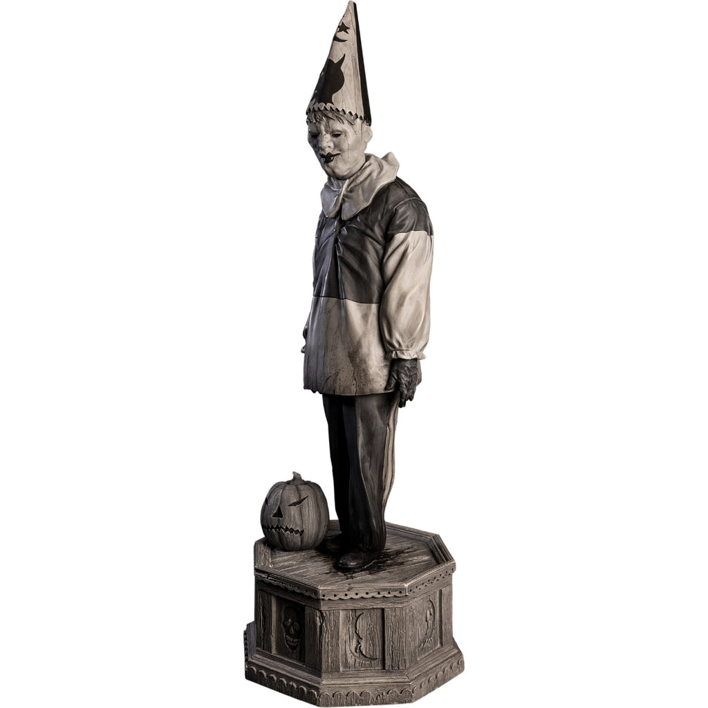 Left side view, statue. Grayscale, Creepy clown, tall pointy hat, black and white shirt with large white collar, black pants, shoes and gloves. Standing next to jack o' lantern on hexagon base made of gravestones