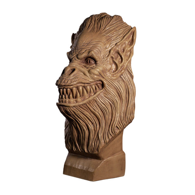 left side view. Crate Beast bust. Clay colored, beast face, wrinkled skin, eyes with vertical long pupils, pointed ears, snout nose, large grinning mouth with many sharp teeth, long hair covering head, lower face and neck. Set on a base.