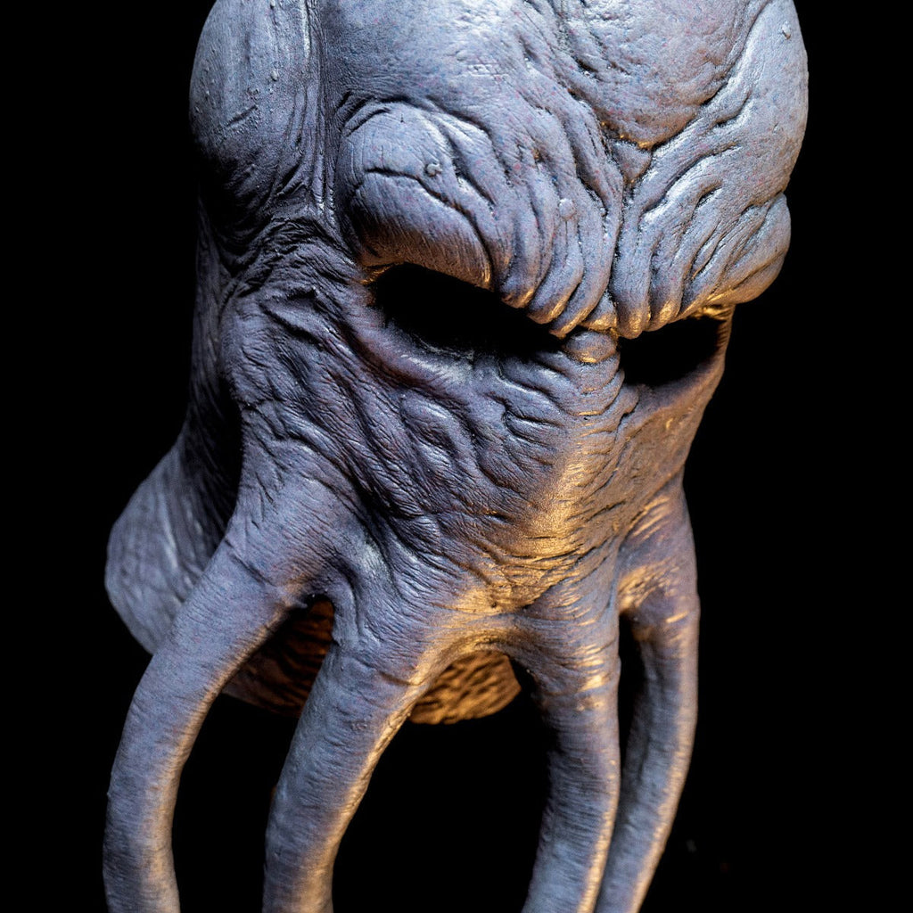 Mask, close up view of face, on black background with dramatic lighting. Lumpy octopus-like head, wrinkled gray flesh. Four long tentacles extending from middle of face.