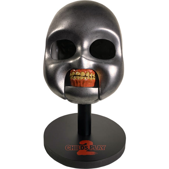 Front view. Good Guys doll skull prop. Metallic silver skull empty black eye sockets. Fleshy red gums with teeth.  Mounted on black wood base, printed red text reads Child's Play 2.