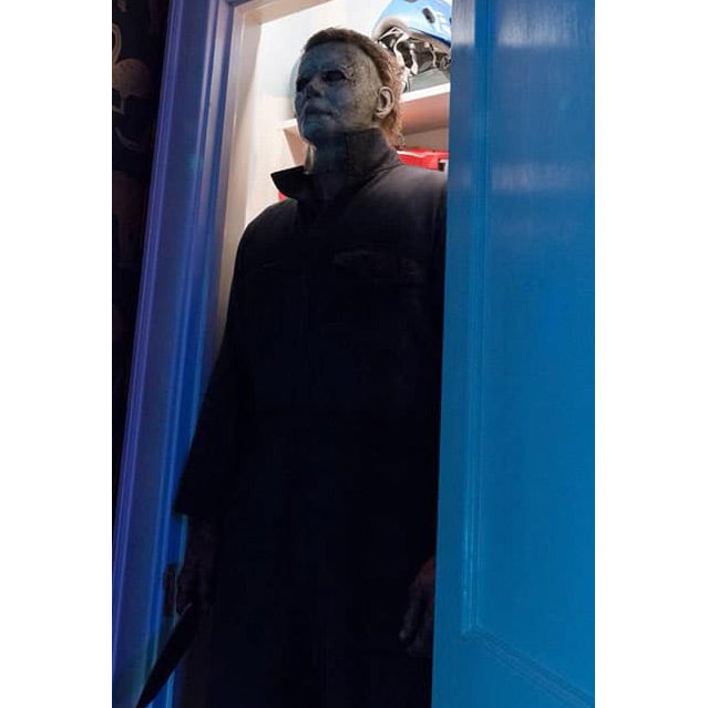 Michael Myers standing in a closet, blue door.  Weathered white mask, dark coveralls, holding knife in right hand.