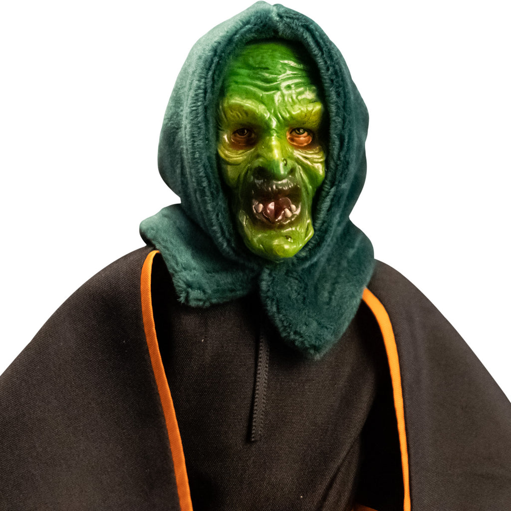 Close up of head and shoulders of witch costume figure. Green witch mask with green hood, wearing black dress and black cape with orange trim.