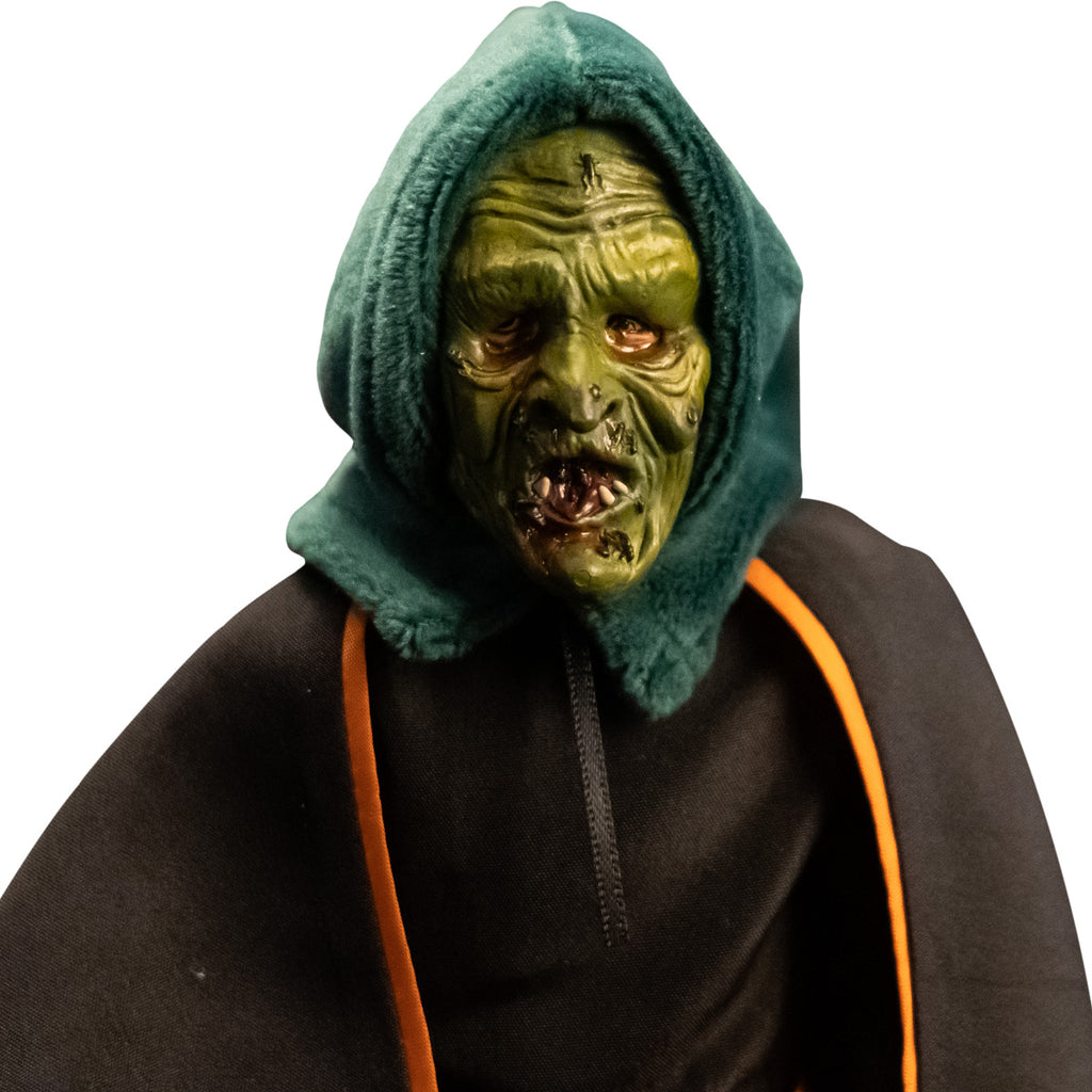 Close up of head and shoulders of witch costume figure. Green rotting witch mask with green hood, wearing black dress and black cape with orange trim.