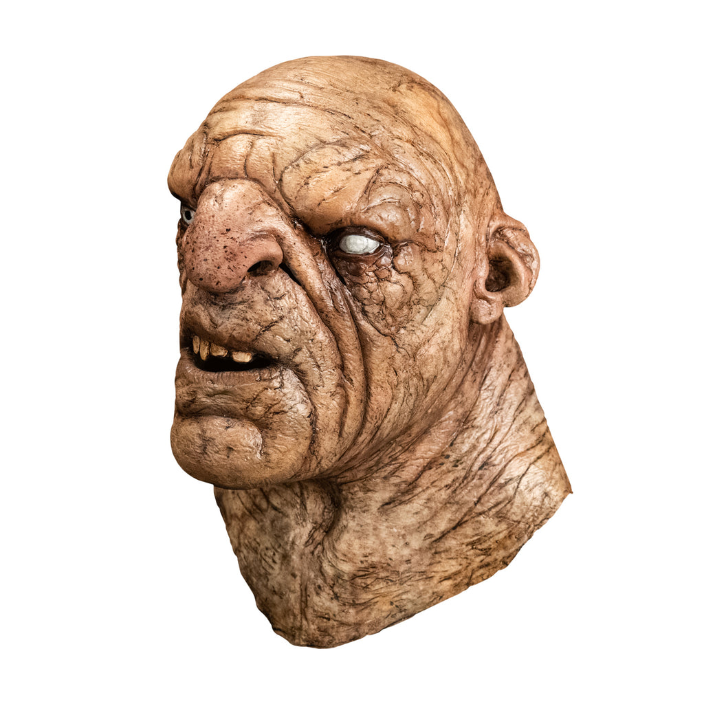 Mask, head and neck, left side view. Troll, bald, wrinkled and dirty tan skin, large nose and chin, mouth open showing teeth. Small ears, right eye blue, left eye white. Thick neck.