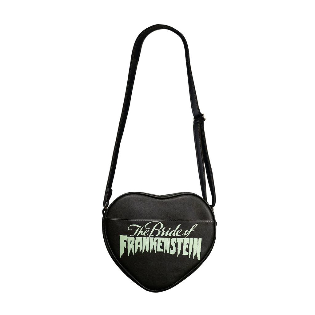 Purse, back view. Black, heart shaped bag, pocket, white text reads The Bride of Frankenstein. Black adjustable strap.