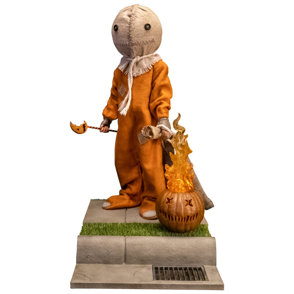 Trick 'r Treat Sam 1/6 scale figure. Head is a stitched burlap sack, Button eyes, stitched mouth, rope tied around neck. wearing orange footy pajamas, brown fingerless gloves. Holding bitten lollipop in right hand, burlap sack in. left hand, flaming jack o' lantern prop in front. Set on sidewalk base, including fake grass, curb and storm drain.