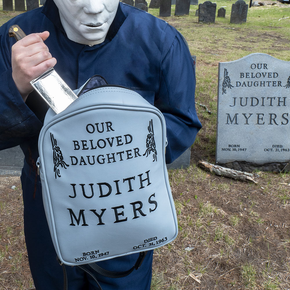 Cemetary location. Person in Michael Myers costume stabbing into top of gray Judith Myers Tombstone bag, Text reads our beloved daughter Judith Myers, born Nov 30 1947, died Oct 31 1963.  Judith Myers tombstone prop in background.