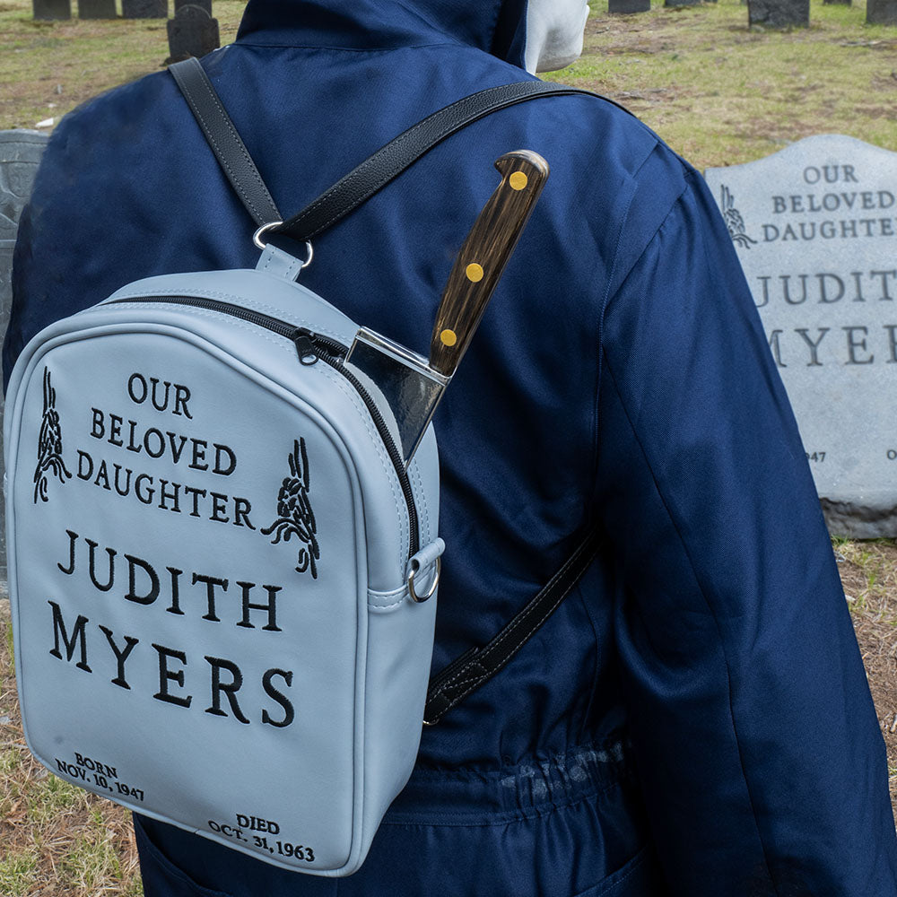 Cemetary location. Person in Michael Myers costume wearing gray Judith Myers Tombstone bag, backpack style with prop knife sticking out the top, Text reads our beloved daughter Judith Myers, born Nov 30 1947, died Oct 31 1963. Judith Myers tombstone prop in background.