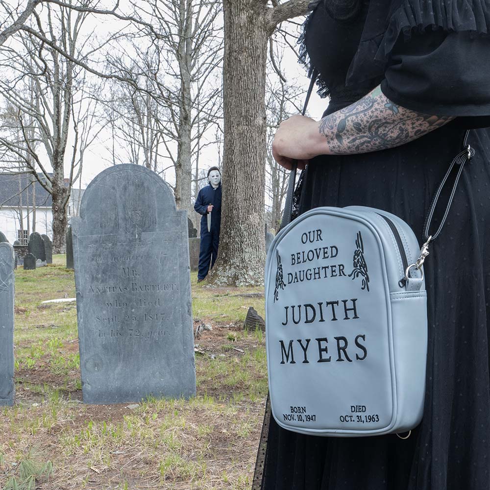 Cemetary location. Woman in black wearing gray Judith Myers Tombstone bag, crossbody style, Text reads our beloved daughter Judith Myers, born Nov 30 1947, died Oct 31 1963. Person in Michael Myers costume holding knife in the background.