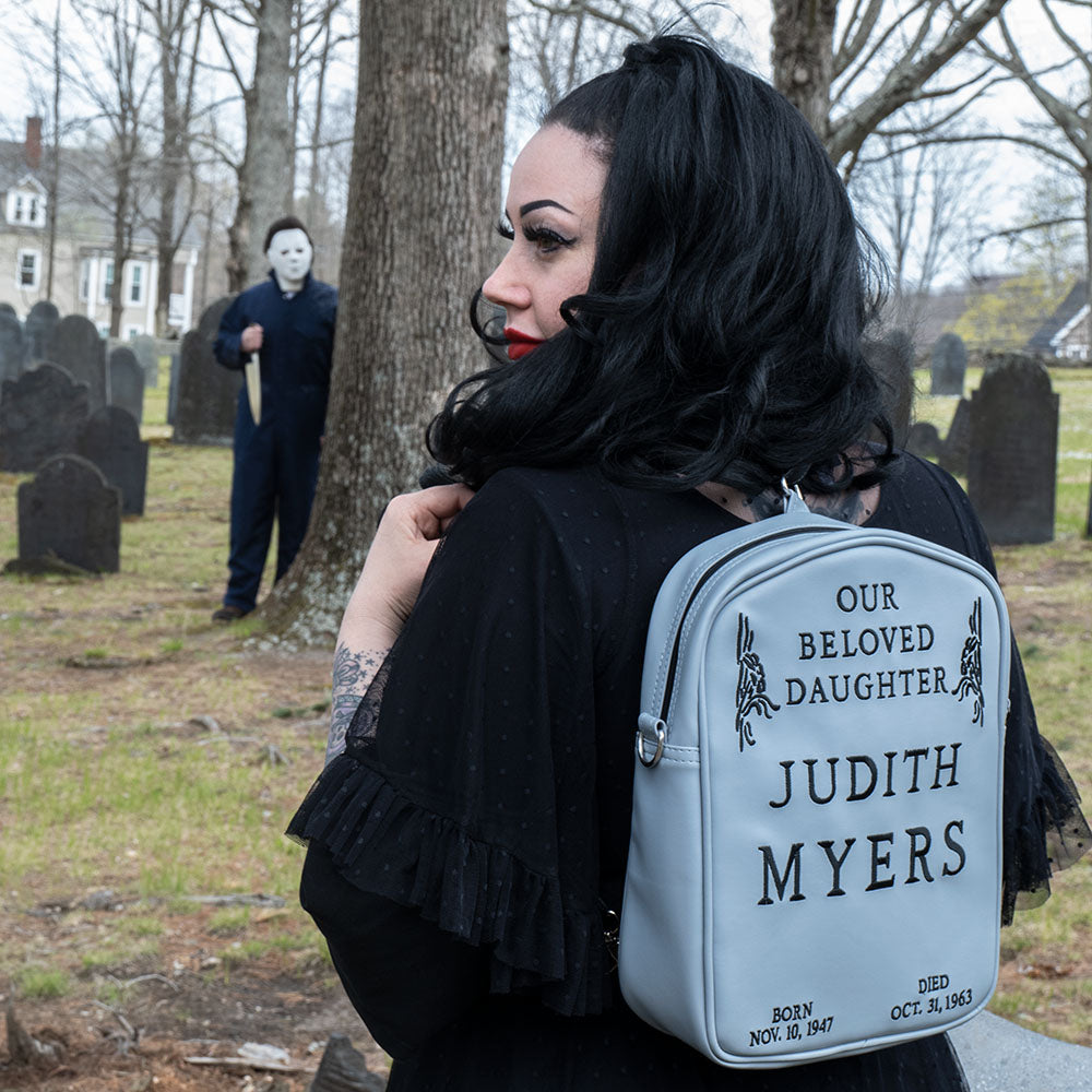 Cemetary location. Woman in black wearing gray Judith Myers Tombstone bag, backpack style. Text reads our beloved daughter Judith Myers, born Nov 30 1947, died Oct 31 1963.  Person in Michael Myers costume holding knife in the background. 