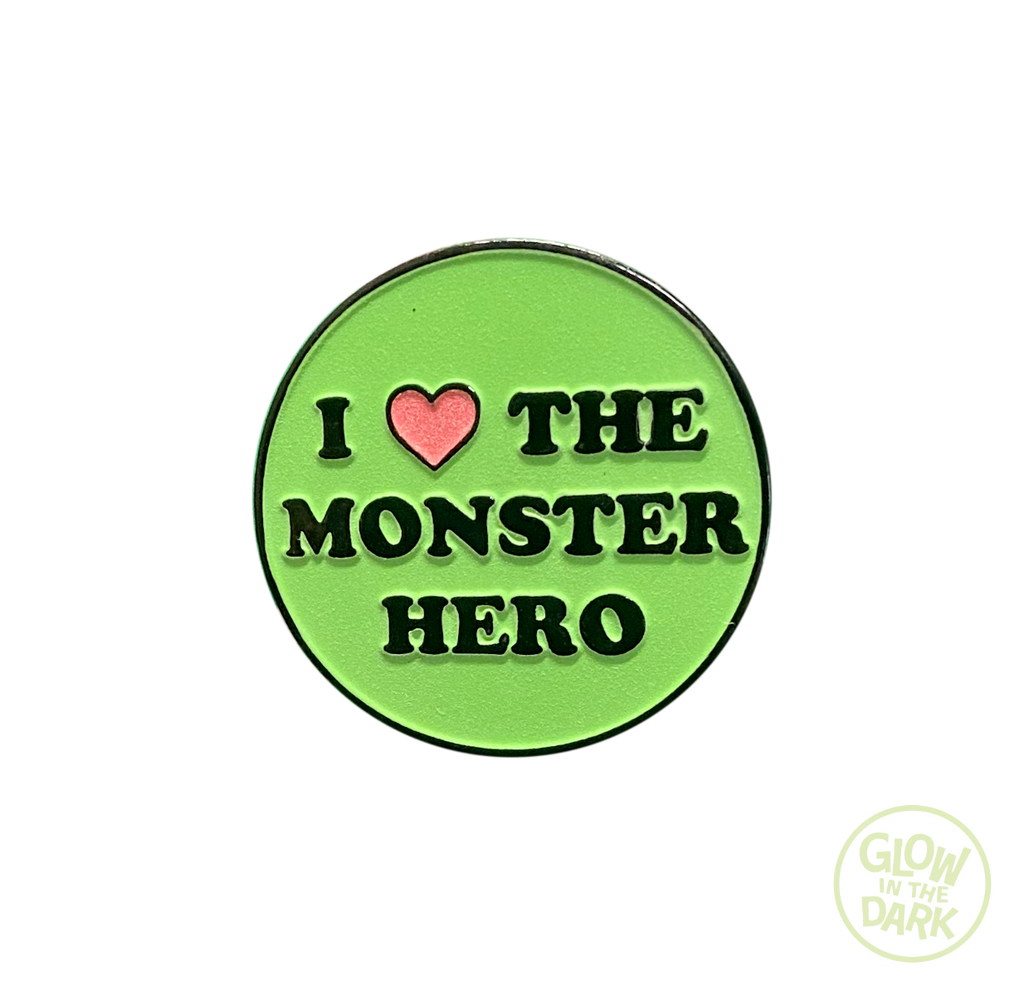 Enamel pin. Green circle, black border.  Black text I. Pink heart, then black text the Monster Hero.  white circle at bottom right of image, text inside reads glow in the dark.