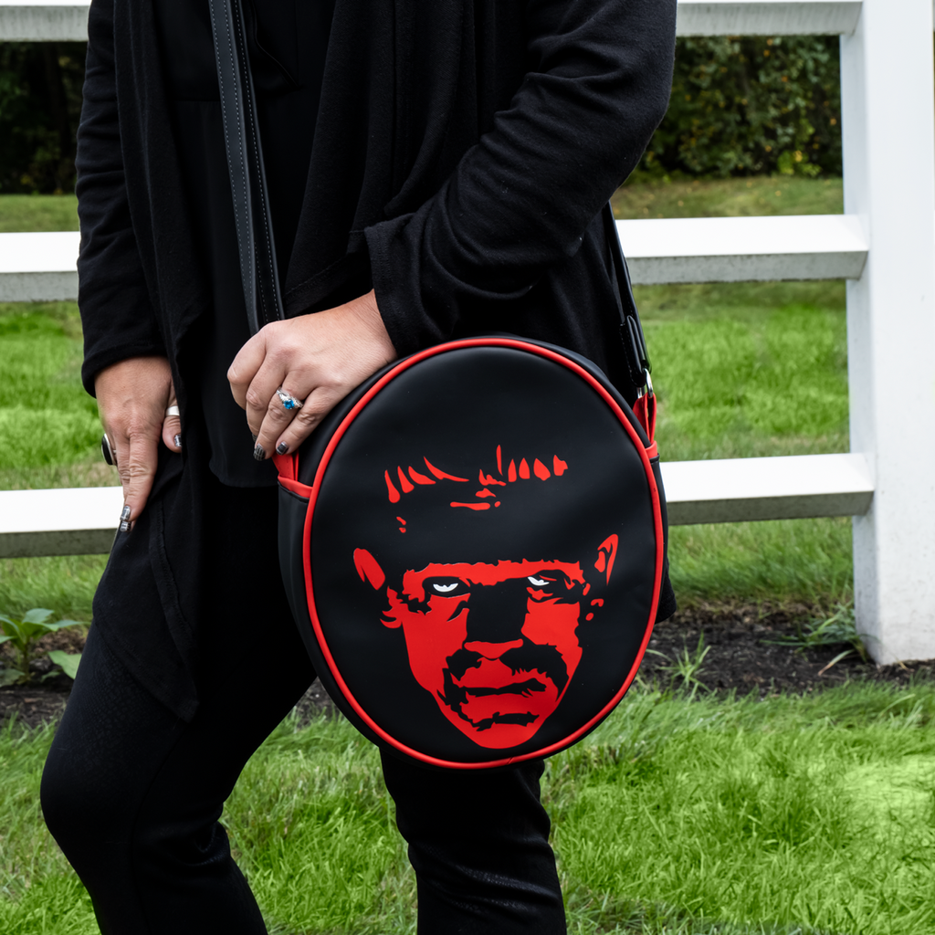 Purse, front view. Worn by person black pants, black jacket.   Black oval, red piping trim, black and red illustration of Frankenstein face, white eyes. Black adjustable strap.