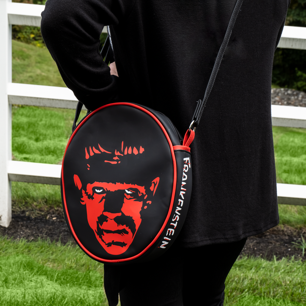 Purse, front and side view. Worn by person black pants, black jacket. Black oval, red piping trim, black and red illustration of Frankenstein face, white eyes. White text on side reads Frankenstein. Black adjustable strap.