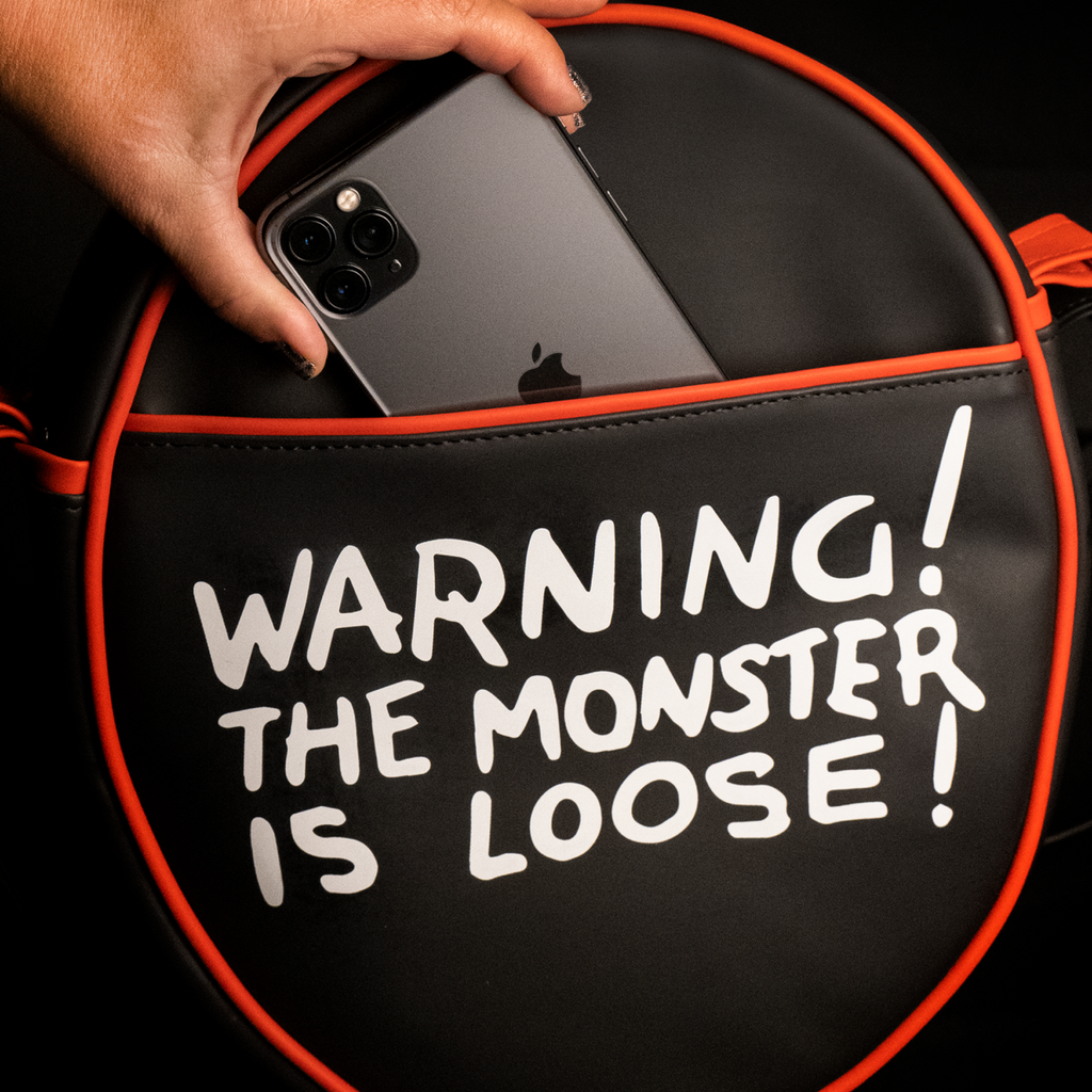 Purse, back view, showing pocket. Black oval, red piping trim, white text reads Warning! The monster is loose!.