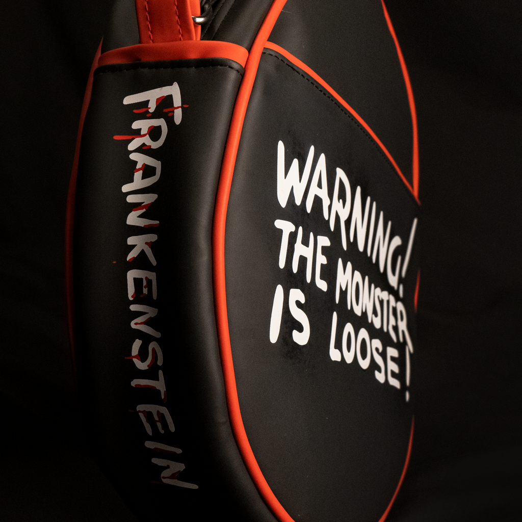 Purse, back and side view, showing pocket. Black oval, red piping trim, white text on side reads Frankenstein, white text on back reads Warning! The monster is loose!.