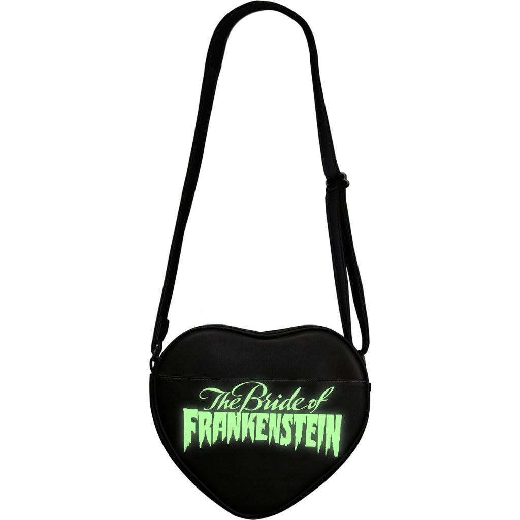 Purse, back view, showing green glow in the dark feature. Black, heart shaped bag, pocket, green glow text reads The Bride of Frankenstein. Black adjustable strap.