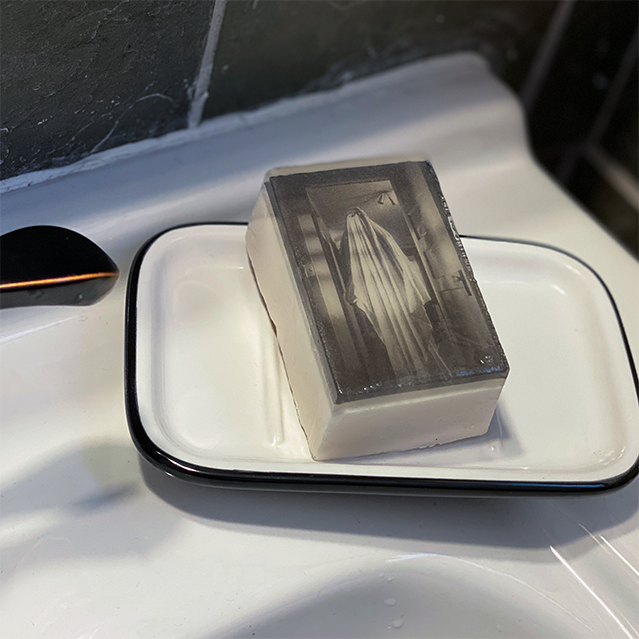 Bar soap. White, with black and white illustration of a sheet ghost standing in a doorway, under clear soap layer. Shown in white and black soap dish on white sink