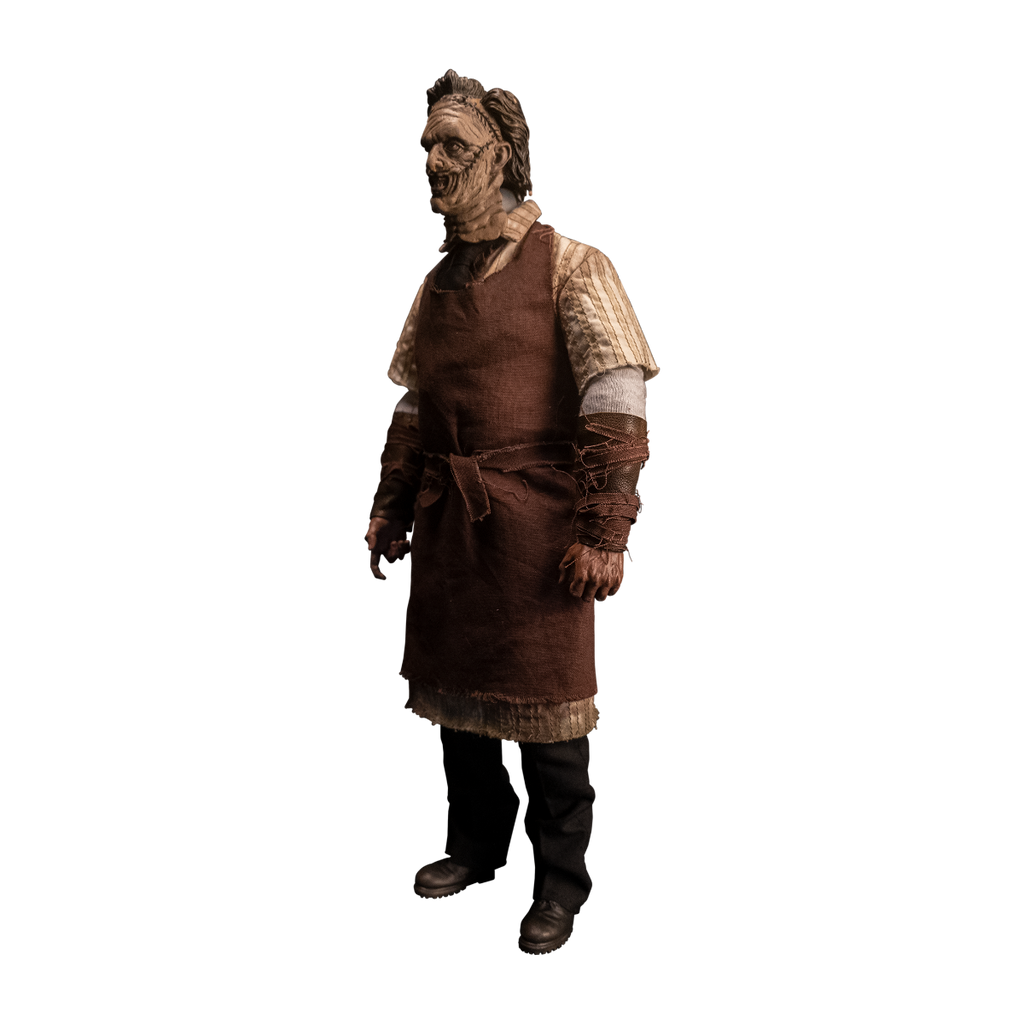 Action figure. Left view. Patchwork mask made of skin, brown hair. Wearing longsleeved white shirt under short sleeved button up shirt, brown tie, brown apron tied at waist. Bracers on forearms, brown strips of leather. Black pants, dark brown shoes.
