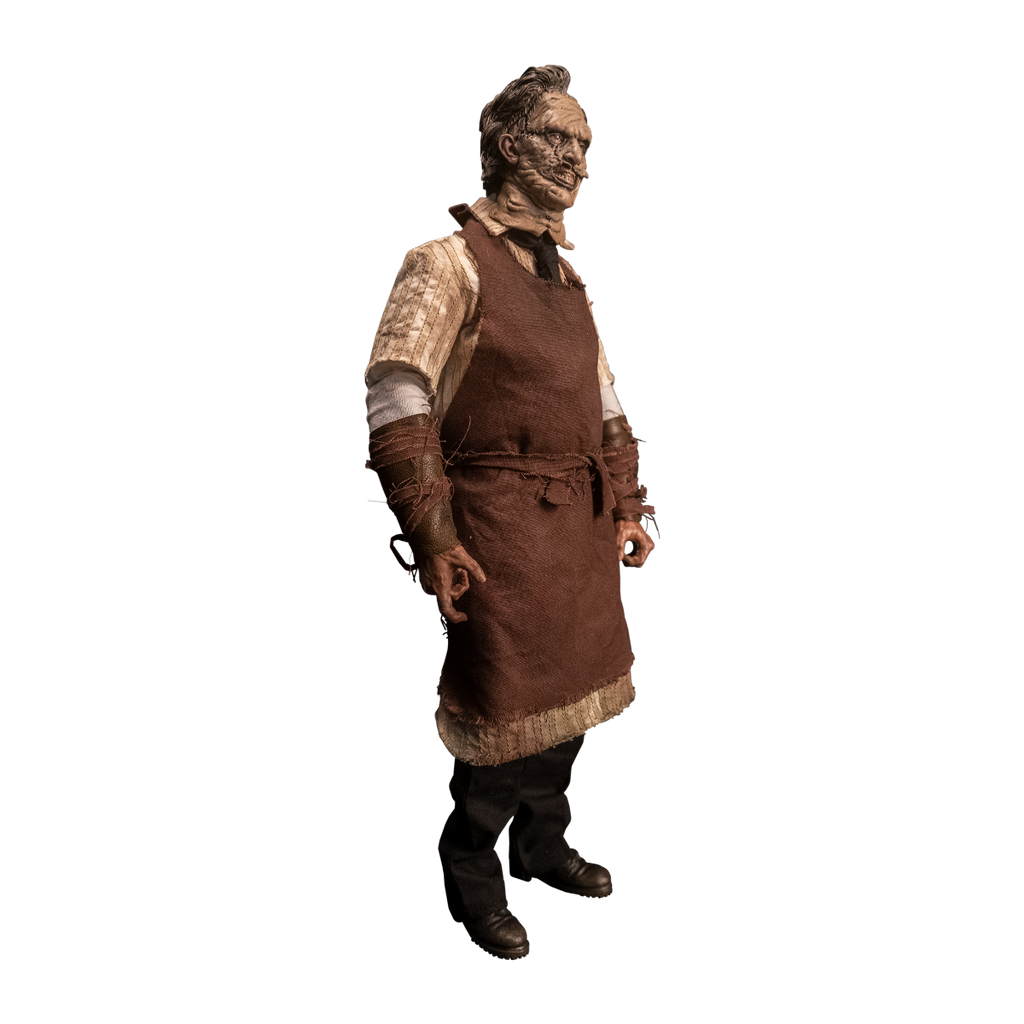 Action figure. Right view. Patchwork mask made of skin, brown hair. Wearing longsleeved white shirt under short sleeved button up shirt, brown tie, brown apron tied at waist. Bracers on forearms, brown strips of leather. Black pants, dark brown shoes.