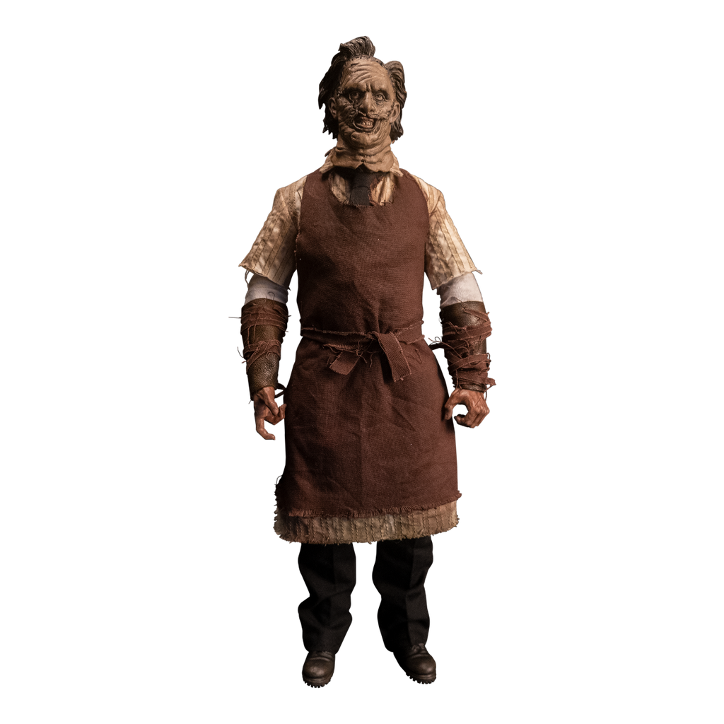 Action figure. Front view. Patchwork mask made of skin, brown hair. Wearing longsleeved white shirt under short sleeved button up shirt, brown tie, brown apron tied at waist. Bracers on forearms, brown strips of leather.  Black pants, dark brown shoes.