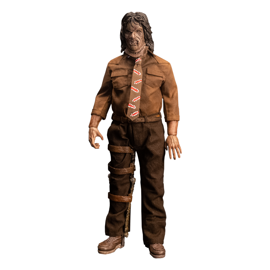 Action figure. Front view. Patchwork mask made of skin, brown hair. Wearing brown collared shirt and brown, red and white necktie, dark brown pants and belt. Brown shoes.  Brown leg brace on right leg.