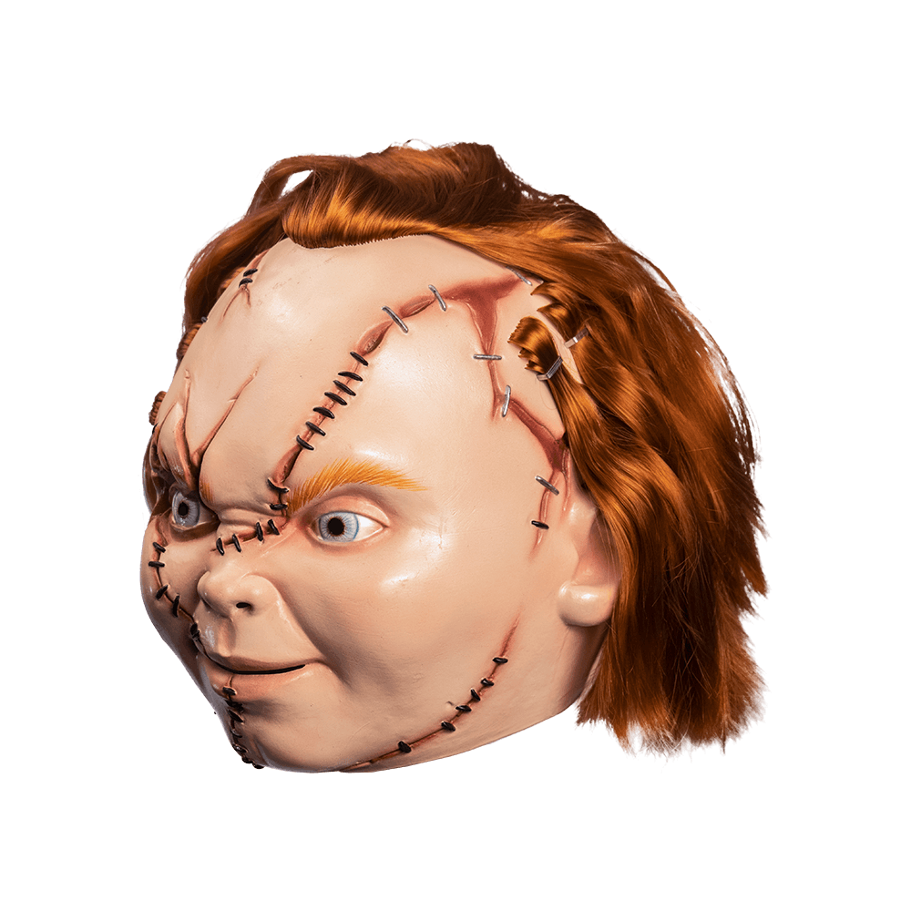 Left side view. Scarred Chucky mask. Red hair, blue eyes, freckles, grinning mouth with teeth showing, cleft chin. Face has many wounds, stitches and staples, sections of hair stapled on.
