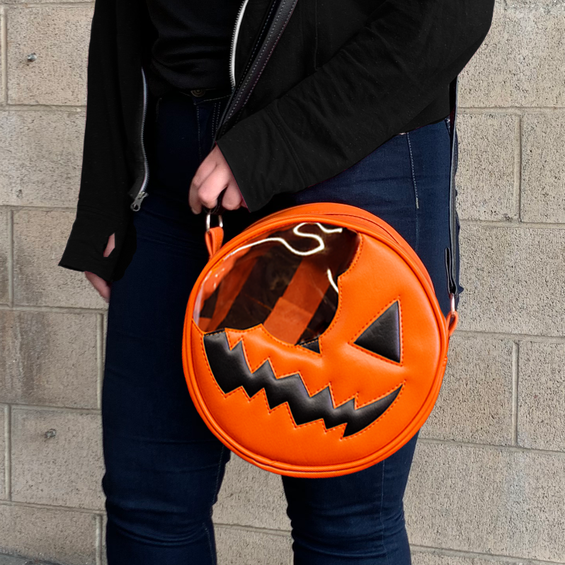 Bag. Front view, being worn by a person in blue jeans, black jacket. Round orange and black, jack o' lantern face appearing to have a bite taken out of the left upper side. Black adjustable strap.