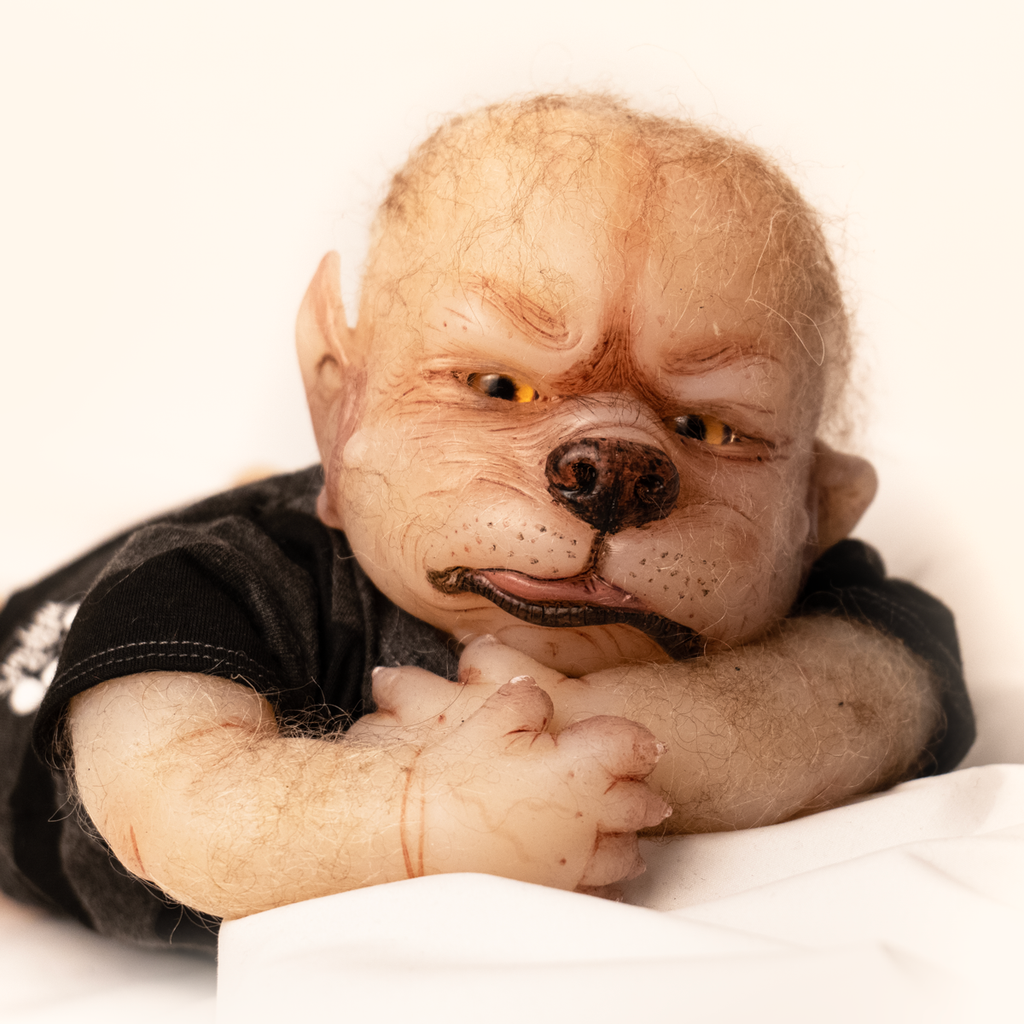 Doll. Upper body view, head shoulders, arms and hands. Tan skin, pointed ears on side of head, sparse light fur on head and arms. Gold eyes, canine nose, mouth with pink tongue slightly sticking out. Paw like hands with claws. Wearing black and gray striped short-sleeved onesie.