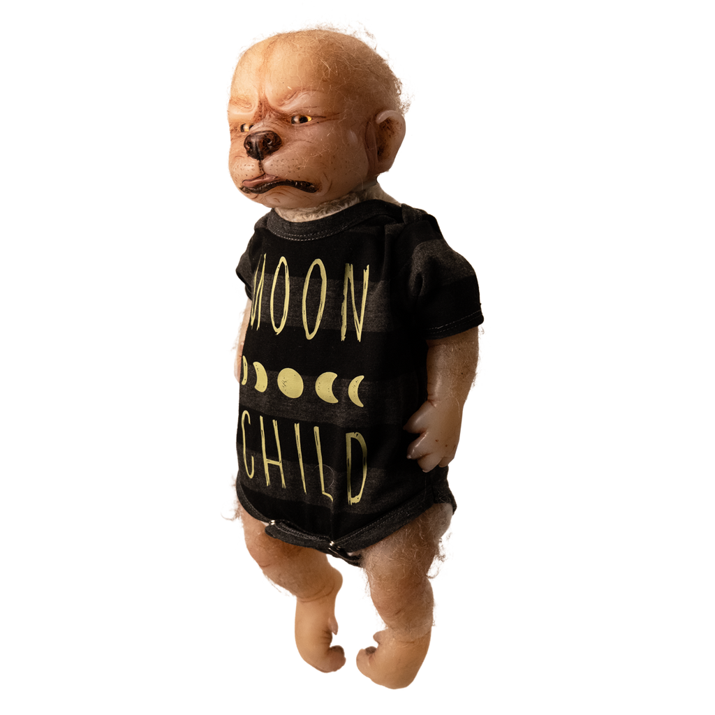 Doll. Full body left view, standing. Tan skin, pointed ears on side of head, sparse light fur on head and arms. Gold eyes, canine nose, mouth with pink tongue slightly sticking out. Paw like hands and feet with claws. Wearing black and gray striped short-sleeved onesie, yellow moon phases, Yellow text on shirt reads Moon Child