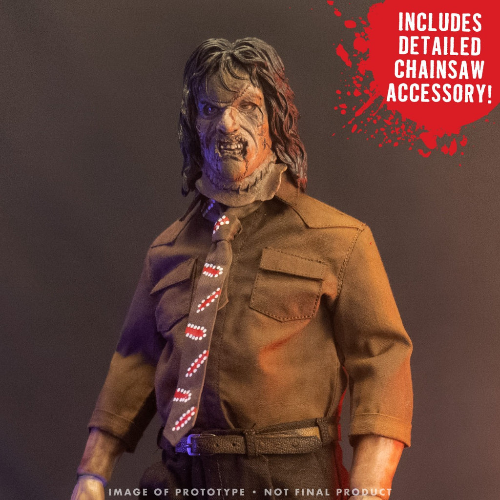Action figure. Closeup, upper body. Patchwork mask made of skin, long brown hair. Wearing brown collared shirt and brown, red and white necktie, dark pants and belt. Red bubble, top right of image, white text reads Includes detailed chainsaw accessory. White text at bottom reads Image of prototype, not final product.