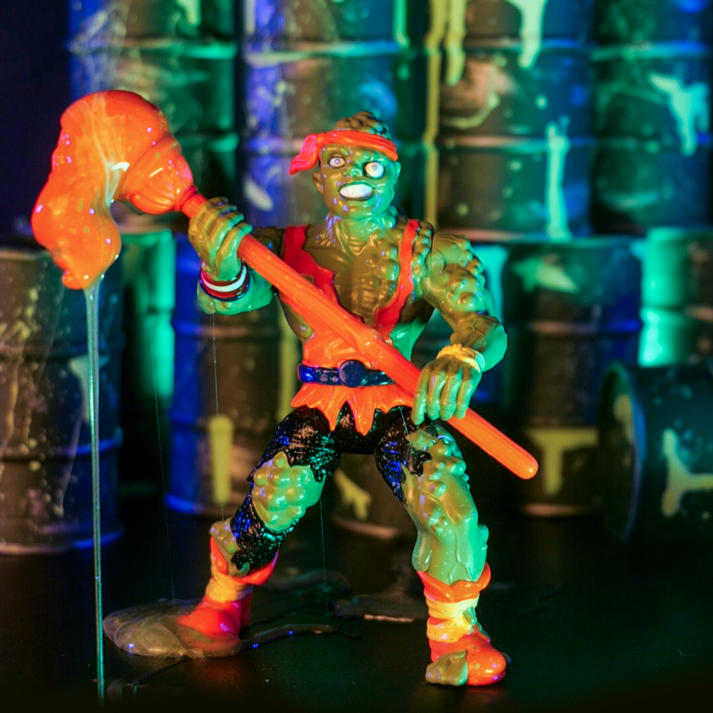 Dramatic lighting, barrels of toxic waste in background. Toxie action figure, front view. Green, lumpy, muscular humanoid figure. Bald, green lumpy blistered flesh, with bright orange headband tied around forehead. Misaligned blue eyes, crooked nose. Lips open showing large teeth. Wearing tattered orange tank top, blue belt, tattered black pants, orange and yellow battered boots. red white and blue wrist band on right wrist, yellow wristband on left wrist. Holding orange mop accessory in both hands.