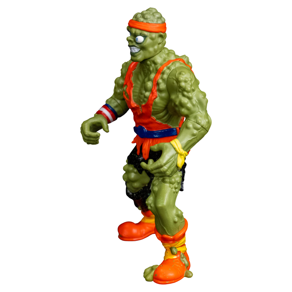 Toxie action figure, left view. Green, lumpy, muscular humanoid figure. Bald, green lumpy blistered flesh, with bright orange headband tied around forehead. Misaligned blue eyes, crooked nose. Lips open showing large teeth. Wearing tattered orange tank top, blue belt, tattered black pants, orange and yellow battered boots. red white and blue wrist band on right wrist, yellow wristband on left wrist.