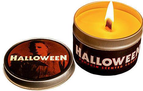 Orange candle in tin, lighted, wide wick.  Lid shows Michael Myers in the background, orange and black, white text reads Halloween.  Candle tin label, black background, white text reads Halloween, orange text reads pumpkin scented candle.