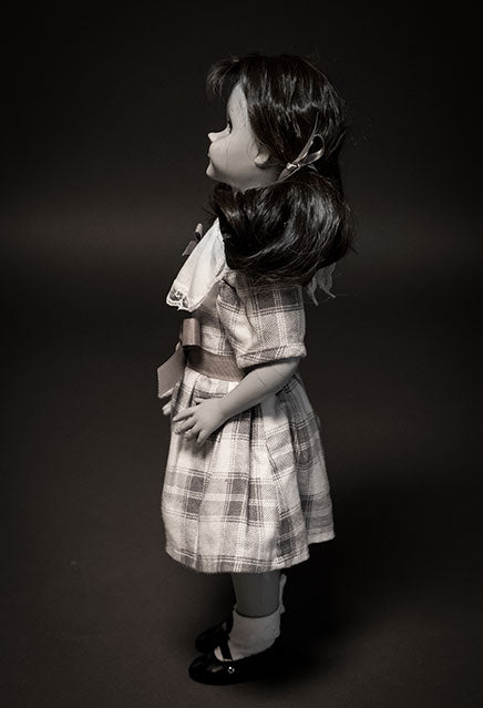 left side view, Black and white, Talky Tina doll wearing plaid dress, with bow at waist, hair in two ponytails with bows. Black shiny dress shoes, white socks.