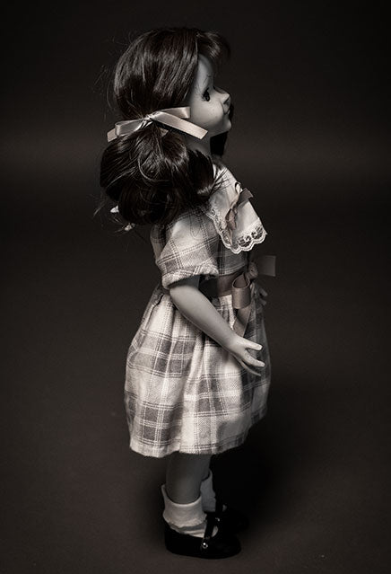 right side view, Black and white, Talky Tina doll wearing plaid dress, with bow at waist, hair in two ponytails with bows. Black shiny dress shoes, white socks.