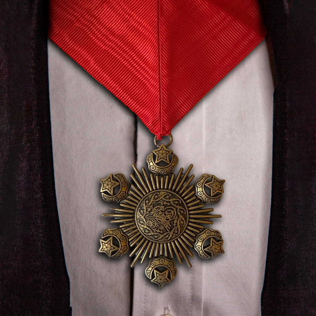 Bronze, hexagonal medallion attached to thick red ribbon. shown on background of white shirt black coat.