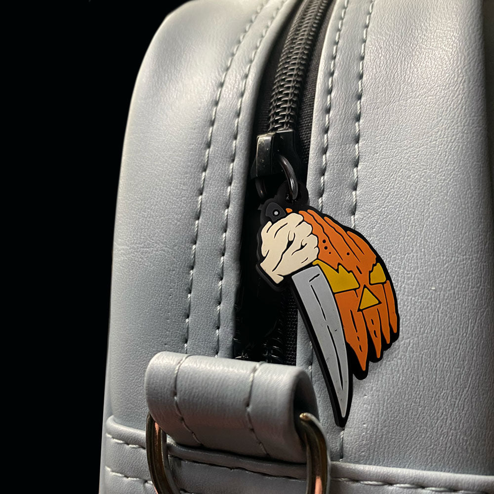 Close up view of zipper pull on gray bag.  Jack o' lantern face and hand holding knife.